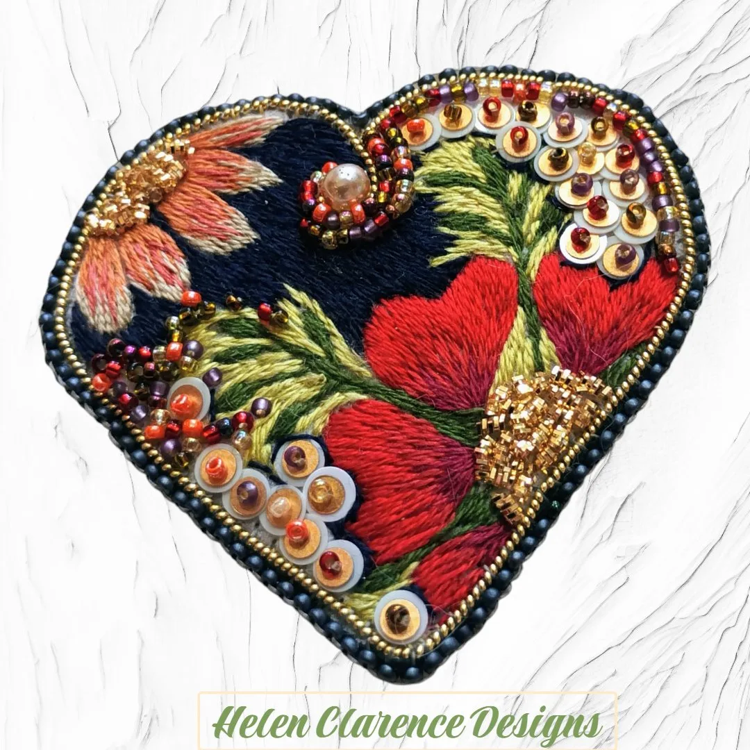 Broche Coeur : Une broche en soie brodée, en forme de cœur, ornée de fleurs, de perles, de paillettes et de fils dorés. Doublée en similicuir suédé.